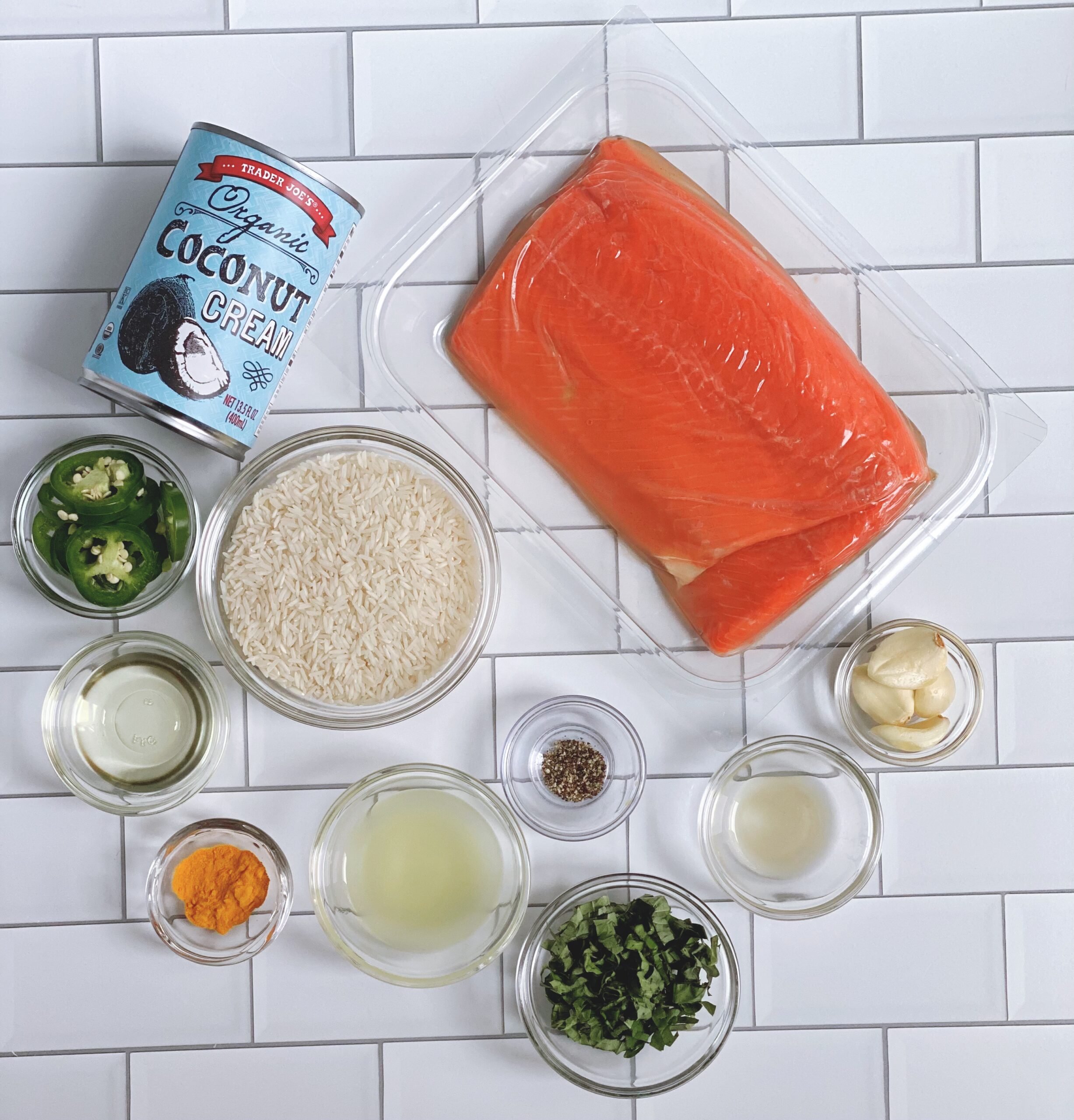 Overhead view of the all the ingredients for the Spicy Coconut Salmon: coconut milk, raw salmon, jasmine rice, jalapenos, tumeric, salt, pepper, scallions, garlic cloves, canola oil and lime juice