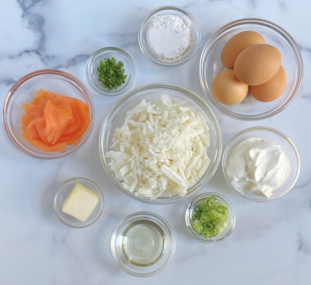 Overhead view of the ingredients for the Potato Cakes with Scrambled Eggs and Smoked Salmon, separated into clear glass bowls, including: shredded frozen potatoes, eggs, sour cream, sliced green onions, canola oil, butter, smoked salmon, finely chopped chives and salt.