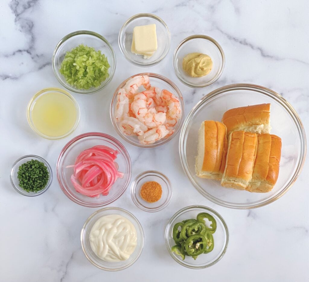 Overhead view of the ingredients needed for the Mini Shrimp Rolls including: Old Bay seasoning, Dijon mustard, diced celery, mayonnaise, chives, pickled jalapenos, shrimp, brioche hot dog rolls, red onions and unsalted butter and lemon juice.