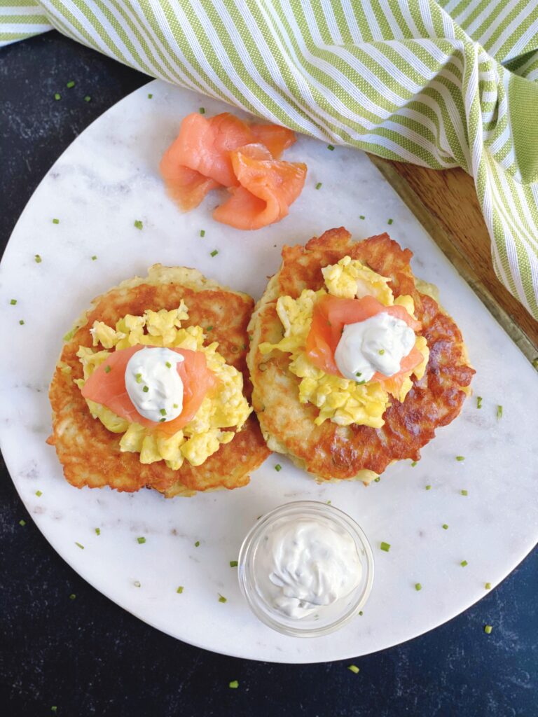 Overhead view of two Potato Cakes with Scrambled Eggs and Smoked Salmon sprinkled with chives and topped with a dallop of sour cream on a white plate.