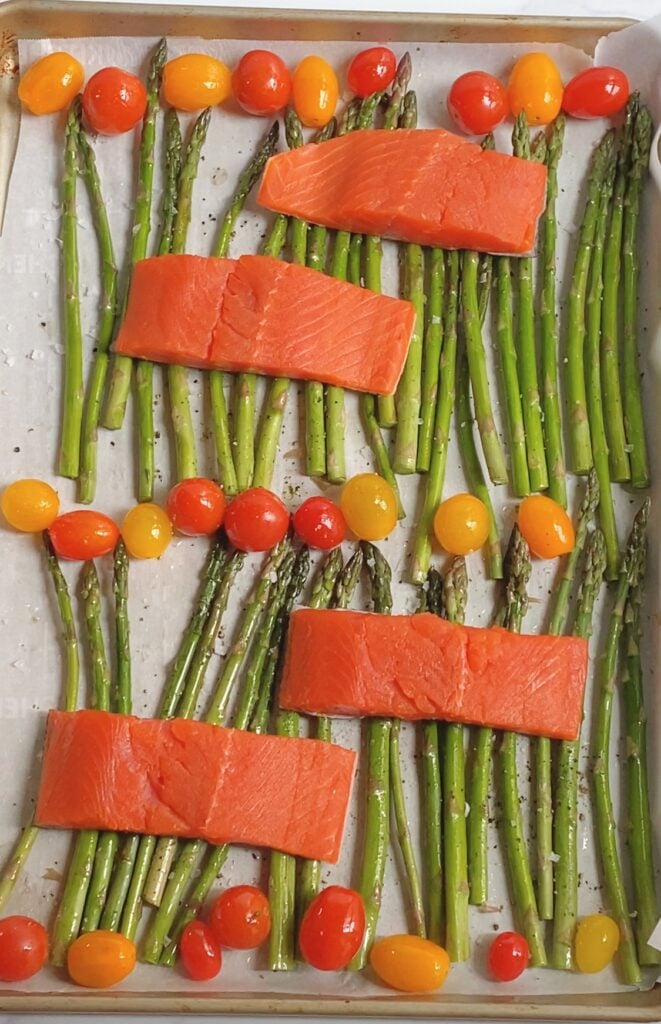 Overhead view of raw salmon filets, cherry tomatoes and asparagus spread out on a sheet pan