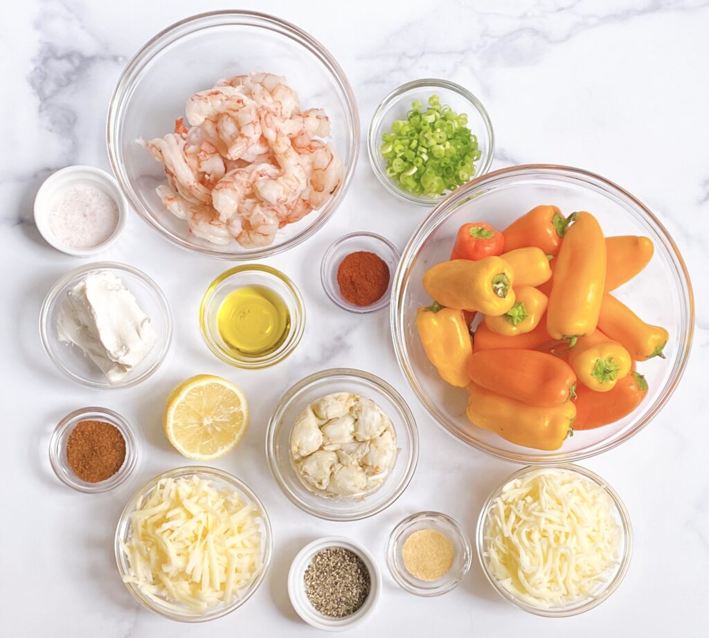 Overhead view of assorted ingredients in separate bowls for Shrimp and Crab Stuffed Mini Sweet Peppers, including: raw shrimp, scallions, mini sweet peppers, shredded white cheddar, garlic powder, salt and pepper, shredded mozzarella, chili powder, softened cream cheese, olive oil, paprika, lump crab and half a lemon