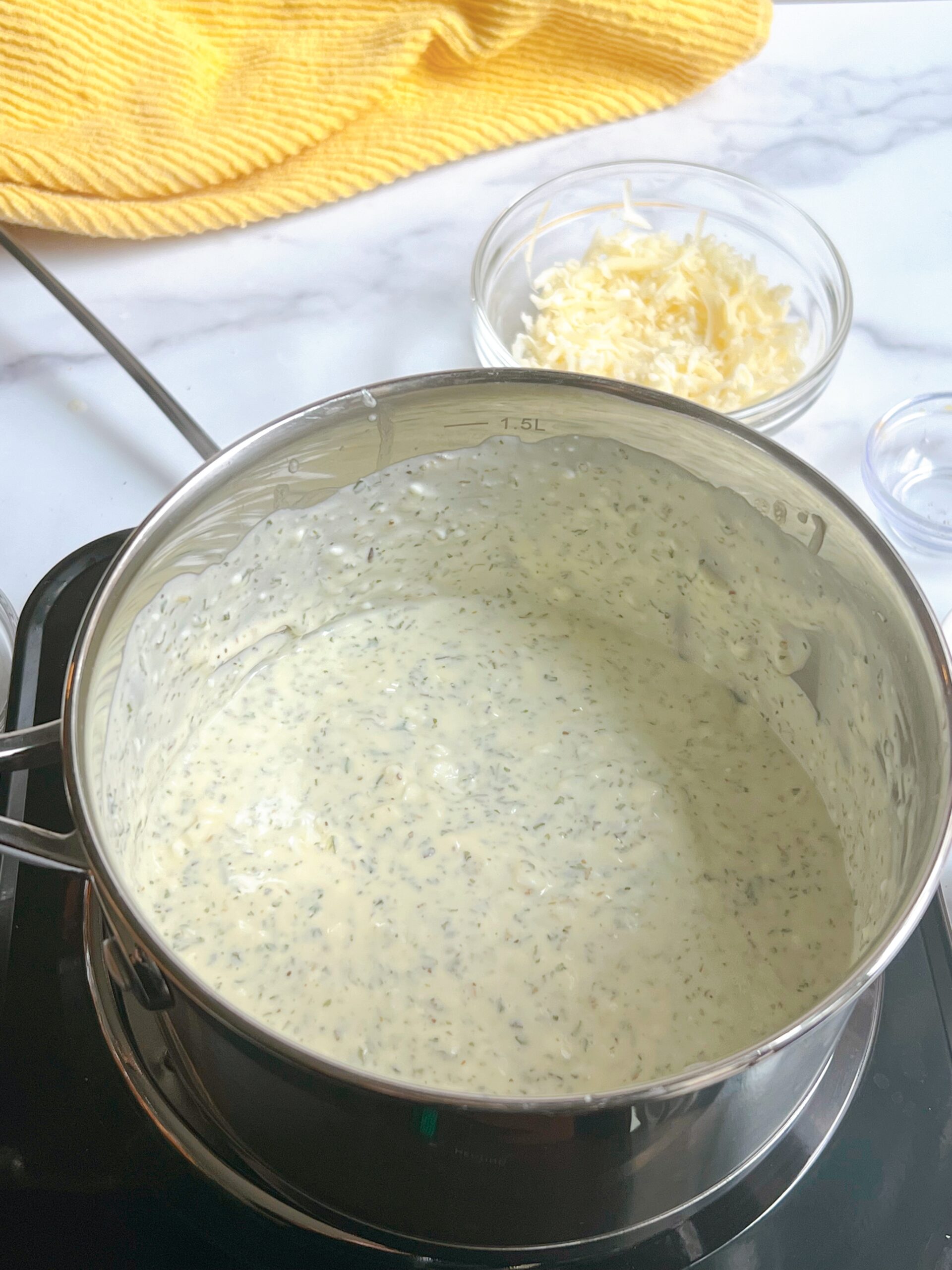 An overhead view of a cream sauce on a burner with visible parsley and tarragon